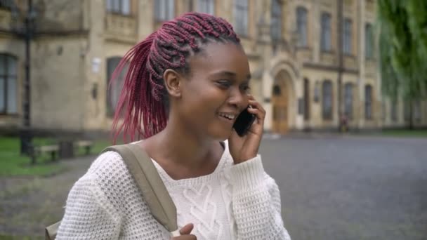 Young african beautiful student talking on phone and laughing, holding backpack and standing in park near university, happy and cheerful - Felvétel, videó
