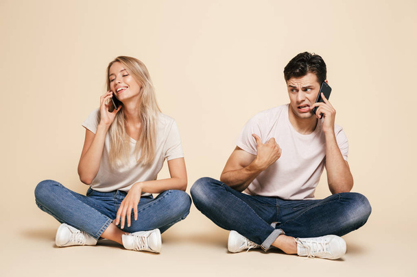 Portrait of a pretty young couple sitting together over beige background, talking on mobile phones - Foto, afbeelding