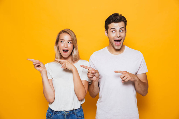 Photo of happy excited young loving couple standing isolated over yellow wall background pointing. - Photo, Image