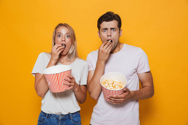 Scared young couple standing isolated over background, eating popcorn - Foto, Imagen