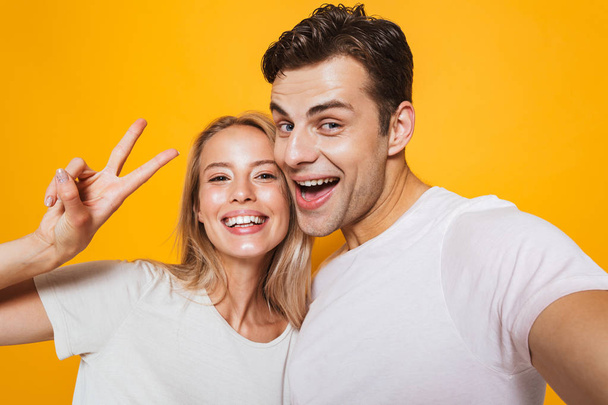Photo of happy excited young loving couple standing isolated over yellow wall background take a selfie by camera showing peace gesture. - Valokuva, kuva