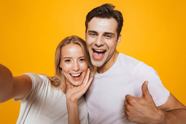 Photo of happy excited young loving couple standing isolated over yellow wall background take a selfie by camera showing thumbs up gesture. - Фото, зображення