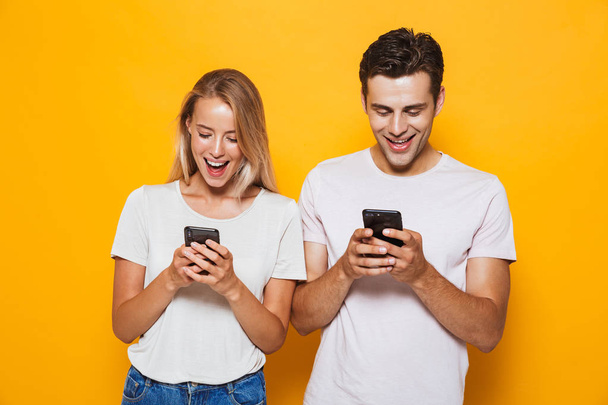 Photo of happy excited young loving couple standing isolated over yellow wall background using mobile phones. - Fotoğraf, Görsel