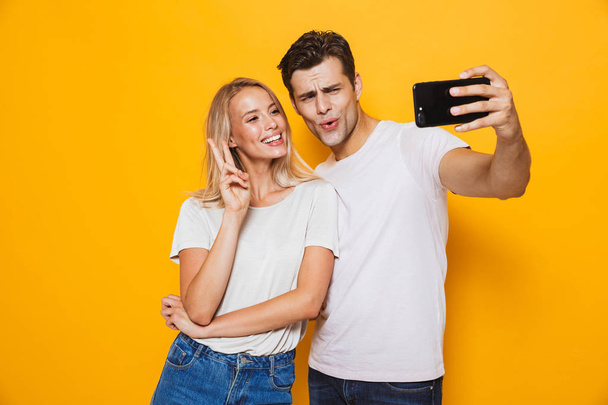 Photo of happy excited young loving couple standing isolated over yellow wall background take a selfie by mobile phone make peace gesture. - Foto, imagen