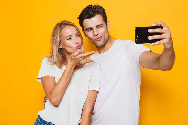 Photo of happy excited young loving couple standing isolated over yellow wall background take a selfie by mobile phone. - Zdjęcie, obraz