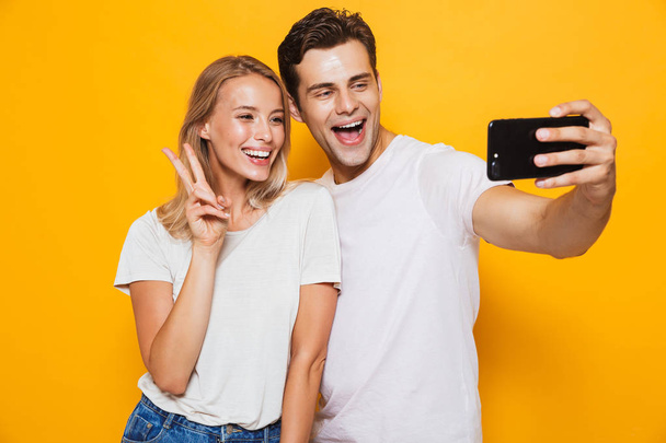 Photo of happy excited young loving couple standing isolated over yellow wall background take a selfie by mobile phone make peace gesture. - Foto, Bild