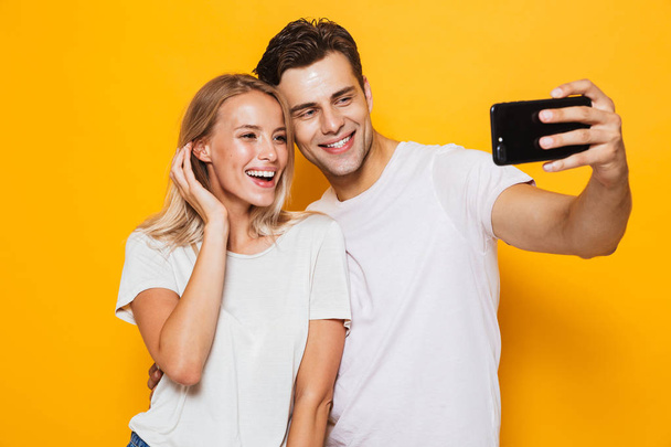 Photo of happy excited young loving couple standing isolated over yellow wall background take a selfie by mobile phone. - Fotografie, Obrázek