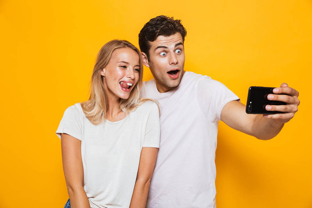 Photo of happy excited young loving couple standing isolated over yellow wall background take a selfie by mobile phone. - Fotoğraf, Görsel