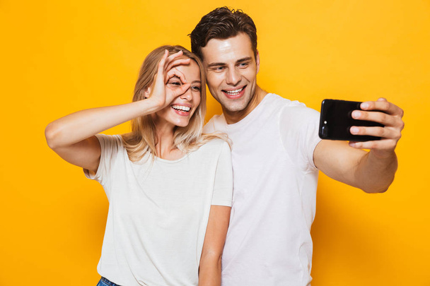 Photo of happy excited young loving couple standing isolated over yellow wall background take a selfie by mobile phone make okay gesture. - Φωτογραφία, εικόνα