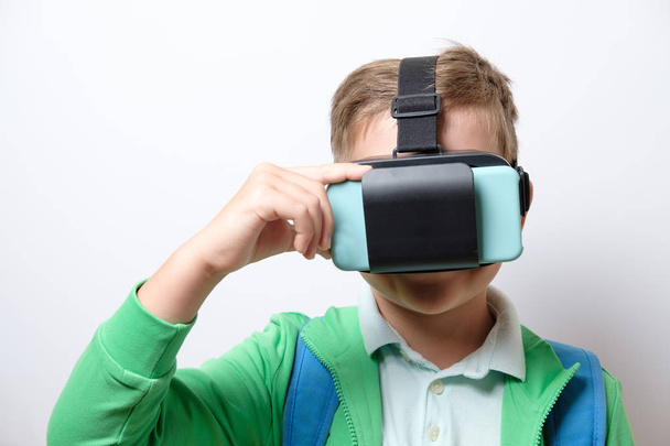 Cute schoolboy with blue backpack wearing virtual reality helmet with arms stretched forward on a white background - Fotó, kép