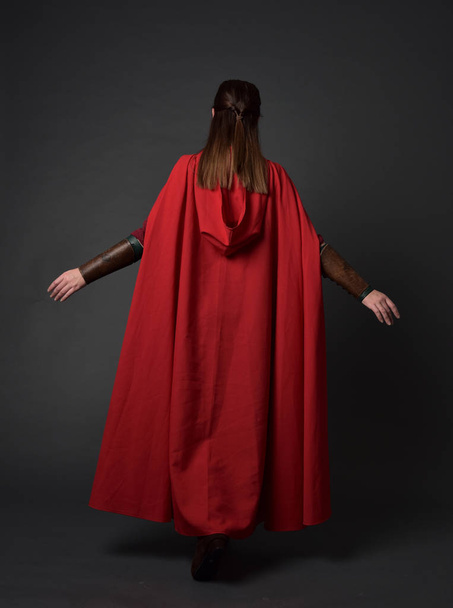 Ganzkörperporträt eines brünetten Mädchens in rotem mittelalterlichen Kostüm und Mantel. Stehpositionen mit dem Rücken zur Kamera auf grauem Studiohintergrund. - Foto, Bild
