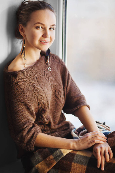vertical portrait of woman sitting on window dressed in brown woolen sweater - Valokuva, kuva