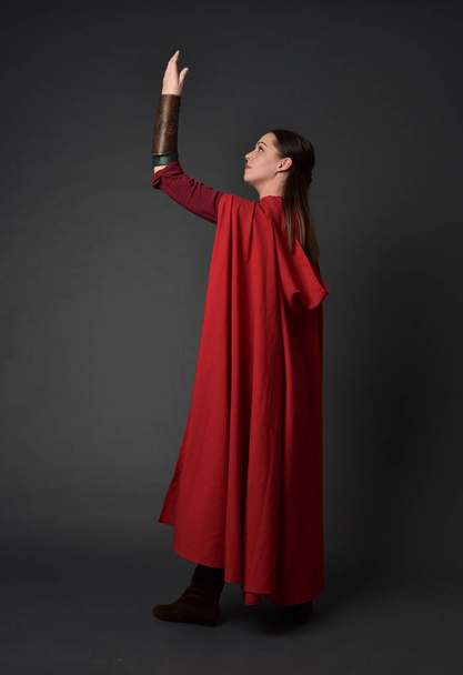 full length portrait of brunette girl wearing red medieval costume and cloak. standing pose  with back to the camera on grey studio background. - Φωτογραφία, εικόνα