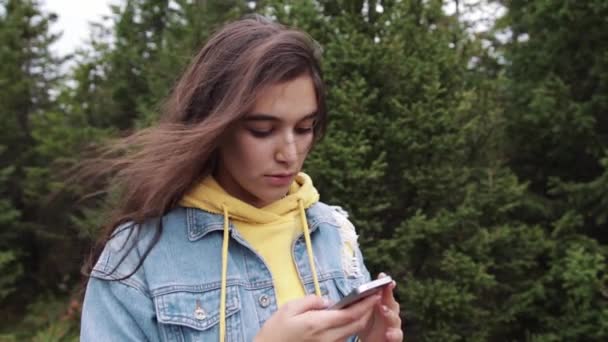 Young teen coed using touch screen smartphone while in nature. Hipster using social media and smiling while reading mobile. Beautiful woman in denim jacket in spruce forest - Metraje, vídeo