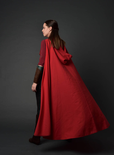 портрет дівчини брюнетки в червоному середньовічному костюмі і плащі. стояча поза ззаду до камери на сірому студійному фоні
. - Фото, зображення