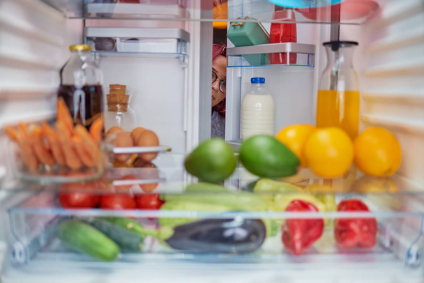 Vrouw gluren in de ontbijtruimte vol boodschappen. Ongezond eten concept. Foto genomen vanaf de binnenkant van de koelkast. - Foto, afbeelding