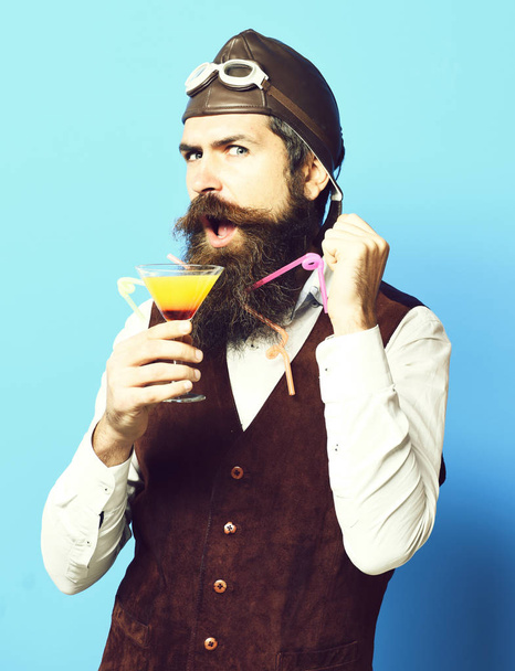 handsome bearded aviator man with long beard and mustache on satisfied face holding glass of alcoholic beverage in vintage suede leather waistcoat with hat and glasses on blue studio background - Фото, зображення