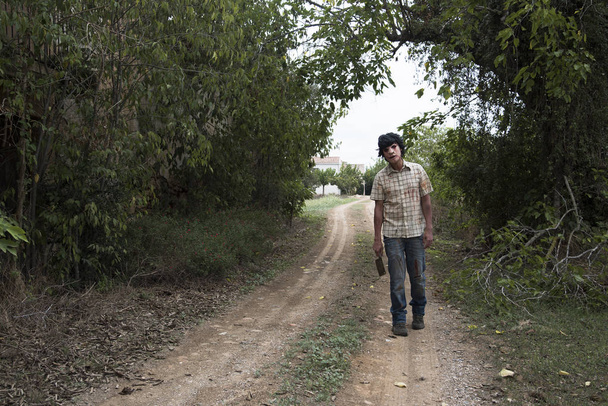 un hombre desfigurado de miedo, vistiendo ropa sucia y andrajosa, con una cuchilla de carne sangrienta y oxidada en la mano, caminando por un camino sucio en un paisaje perturbador
 - Foto, Imagen