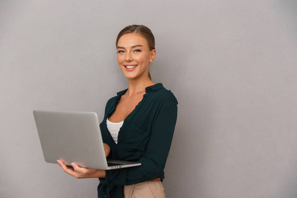 Confident young businesswoman standing over gray background, working on laptop computer - Foto, Imagem