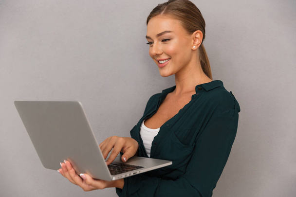 Happy young businesswoman standing over gray background, working on laptop computer - Foto, imagen