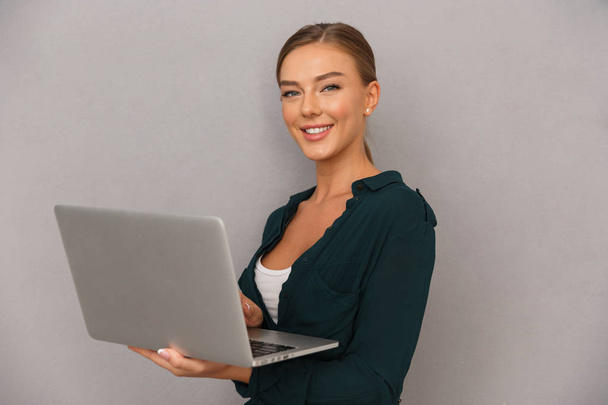Pretty young businesswoman standing over gray background, working on laptop computer - Zdjęcie, obraz