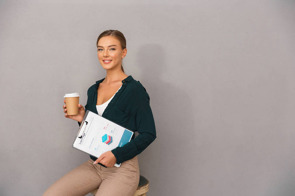 Smiling young businesswoman over gray background, holding documents, sitting, drinking coffee - Fotografie, Obrázek