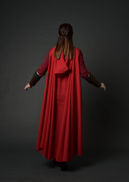 Portret van de volledige lengte van brunette meisje dragen rode middeleeuws kostuum en mantel. staande poseren met de rug naar de camera op grijs studio achtergrond. - Foto, afbeelding