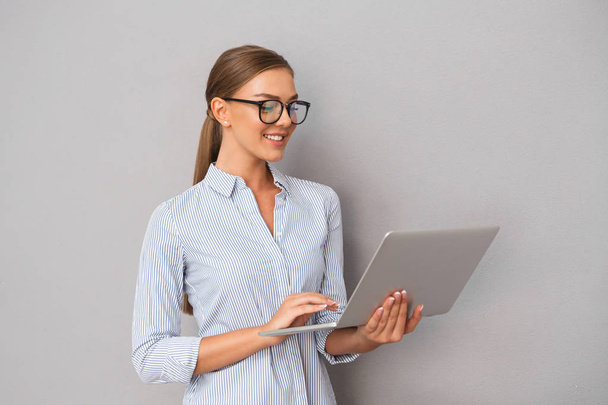 Image of beautiful smiling business woman standing over grey wall background using laptop computer. - Φωτογραφία, εικόνα