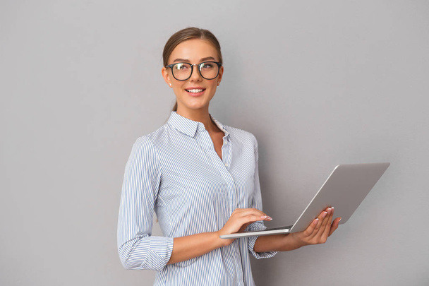 Imagen de una hermosa mujer de negocios sonriente de pie sobre fondo de pared gris usando computadora portátil
. - Foto, imagen
