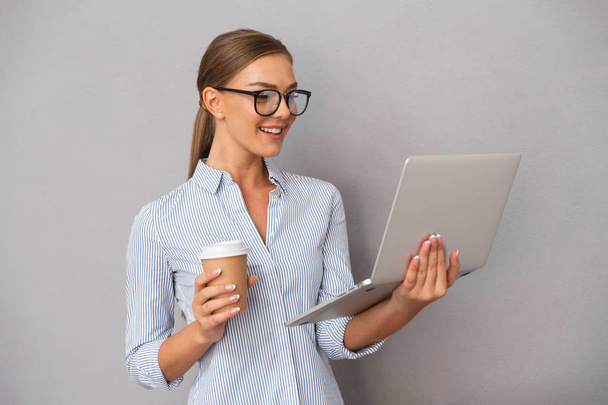 Cheerful young businesswoman standing over gray background, working on laptop computer, drinking coffee - Fotografie, Obrázek