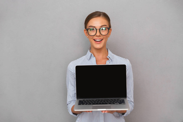Happy young businesswoman standing over gray background, showing blank screen laptop computer - Фото, зображення