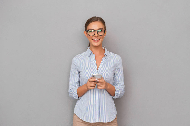 Image of beautiful smiling business woman standing over grey wall background using mobile phone. - Photo, Image