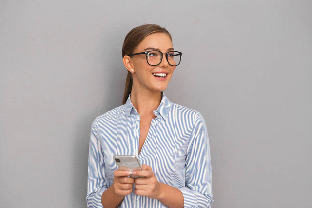 Image of beautiful smiling business woman standing over grey wall background using mobile phone. - Φωτογραφία, εικόνα