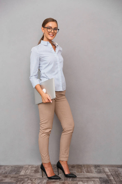 Full length of a smiling young businesswoman standing over gray background, carrying laptop computer - Fotografie, Obrázek