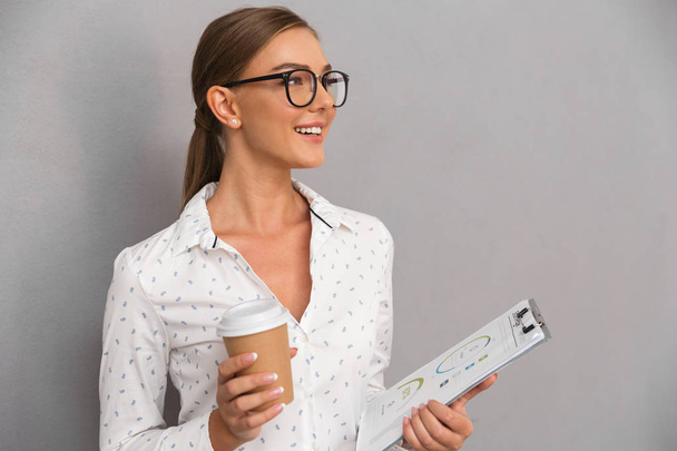 Image of beautiful business woman standing over grey wall background holding clipboard drinking coffee. - Valokuva, kuva
