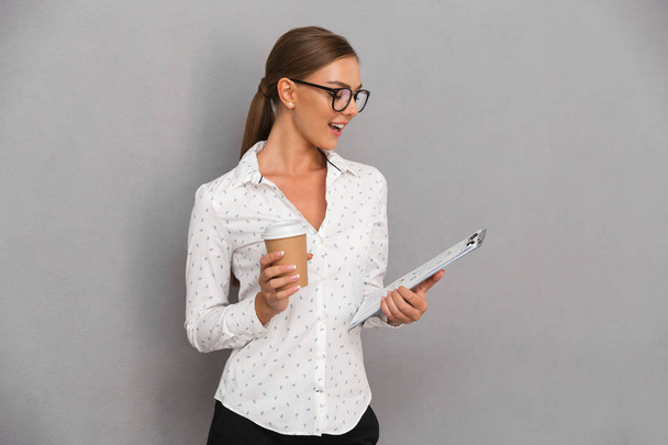 Image of beautiful business woman standing over grey wall background holding clipboard drinking coffee. - Zdjęcie, obraz