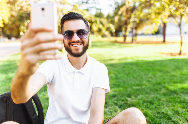 stylish hipster student in a white shirt sitting on the lawn in the Park and takes a selfie on your smartphone - Foto, afbeelding