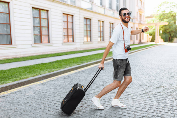 turista positivo com barba, com mala e câmera, correndo pelas ruas, em direção ao descanso
 - Foto, Imagem