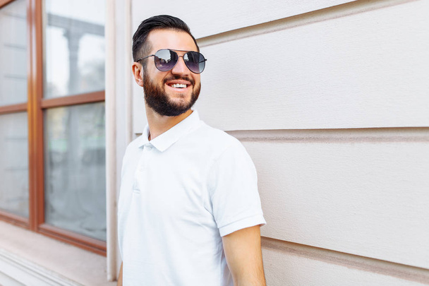Stylish hipster in a white t-shirt with sunglasses and a beard, standing near a white wooden wall - Fotografie, Obrázek