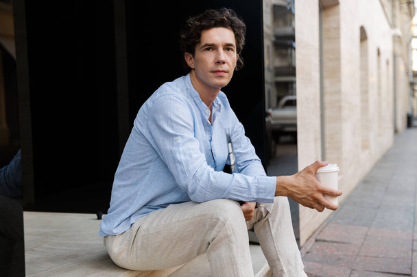 Handsome young casual man drinking coffee while sitting at the city street - Φωτογραφία, εικόνα