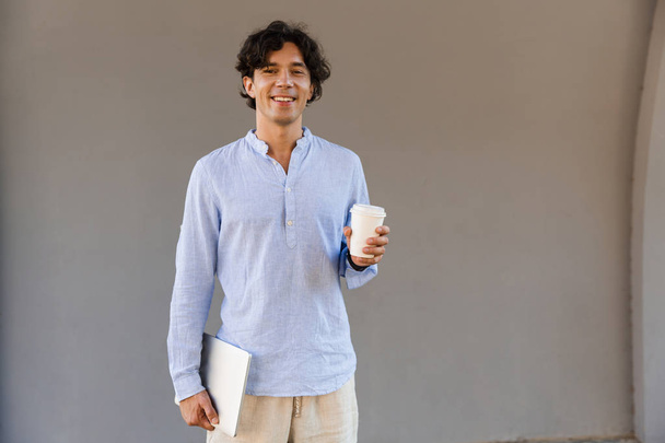 Smiling man dressed in shirt drinking coffee while standing outdoors at the city, holding laptop - Φωτογραφία, εικόνα