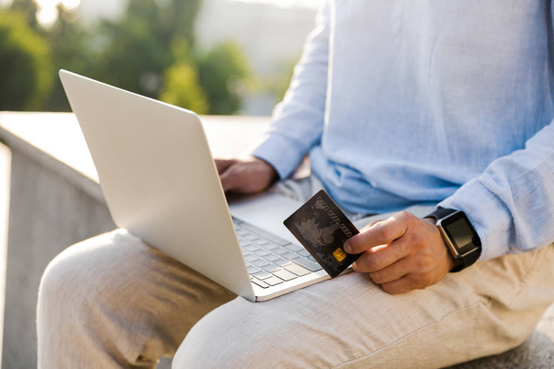 Close up of a man holding plastic credit card while using laptop outdoors - Foto, imagen