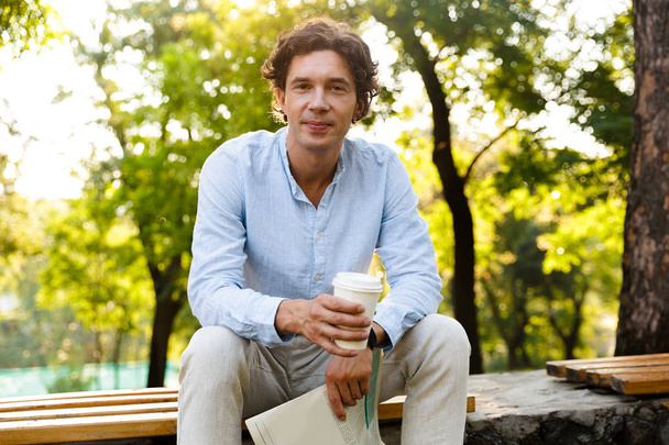 Attractive young casual man holding newspaper while sitting at the city park, drinking coffee - 写真・画像