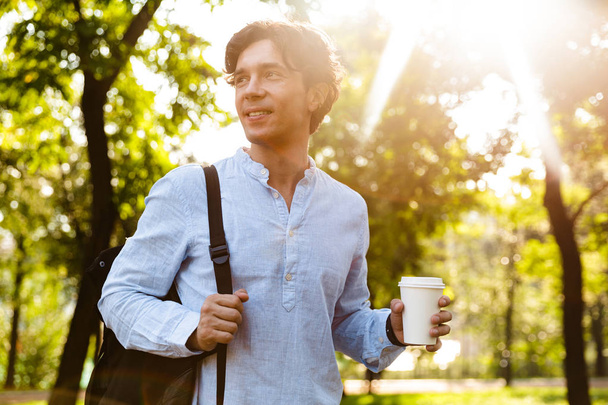Smiling young casual man drinking coffee while walking at the city park, carrying backpack - 写真・画像