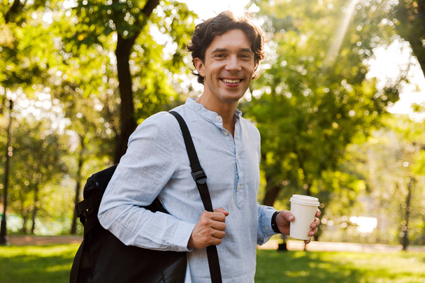 Happy young casual man drinking coffee while walking at the city park, carrying backpack - Фото, изображение