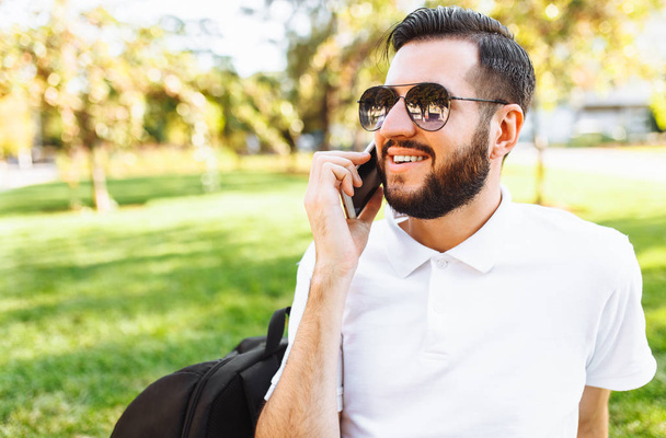 Business and stylish hipster in a white shirt and sunglasses, sitting on the lawn in the Park and talking on the phone - Photo, image