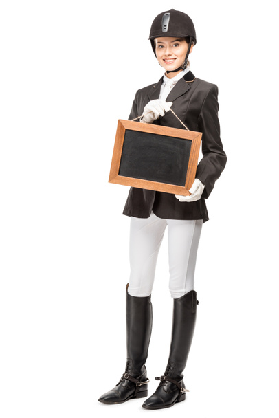 beautiful young horsewoman in uniform holding blank chalkboard isolated on white - Photo, Image