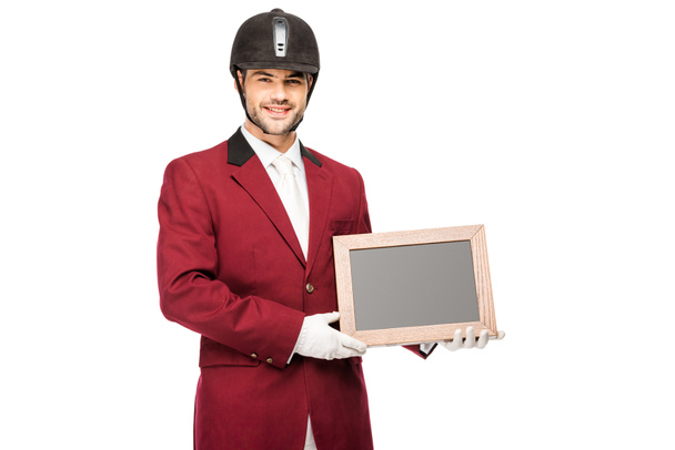 happy young horseman in uniform holding blank chalkboard and looking at camera isolated on white - Foto, imagen