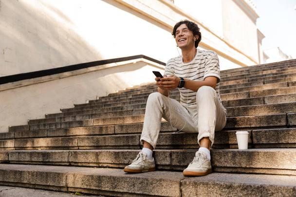 Image of handsome young man sitting on steps outdoors listening music using mobile phone. - Foto, immagini