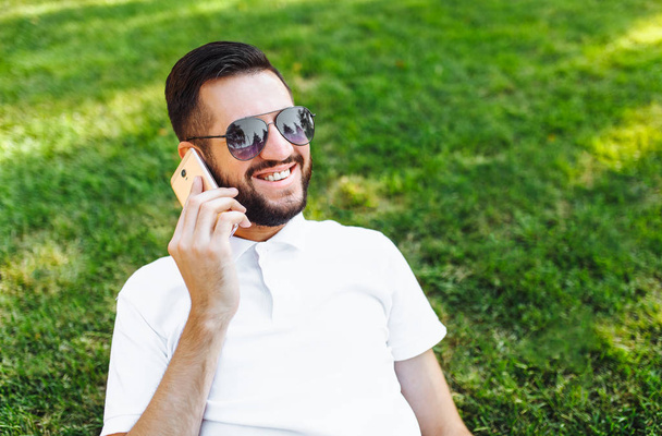 Business and stylish hipster in a white shirt and sunglasses, sitting on the lawn in the Park and talking on the phone - Фото, изображение